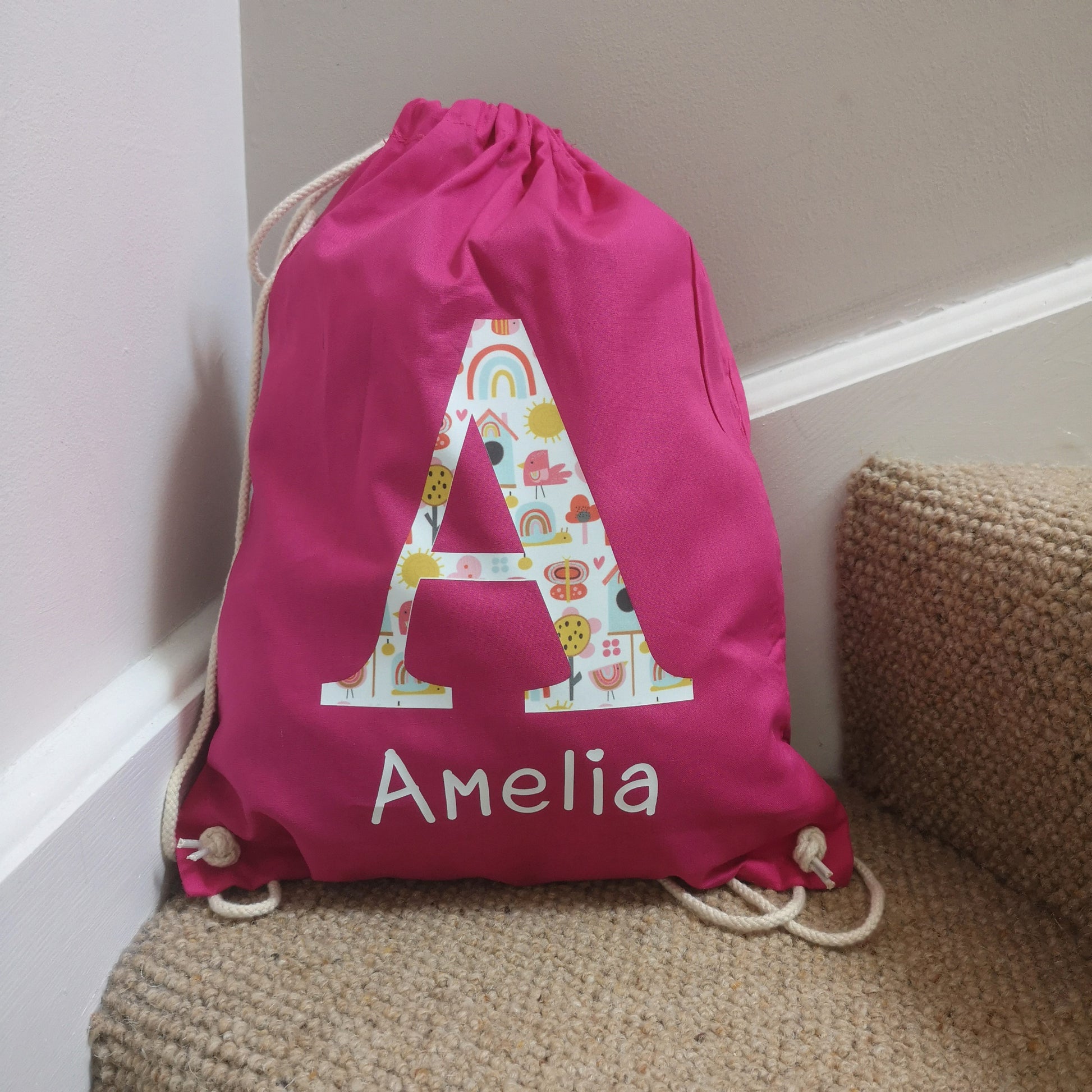 A fuschia pink personalised light drawstring bag with a patterned Initial and their name underneath