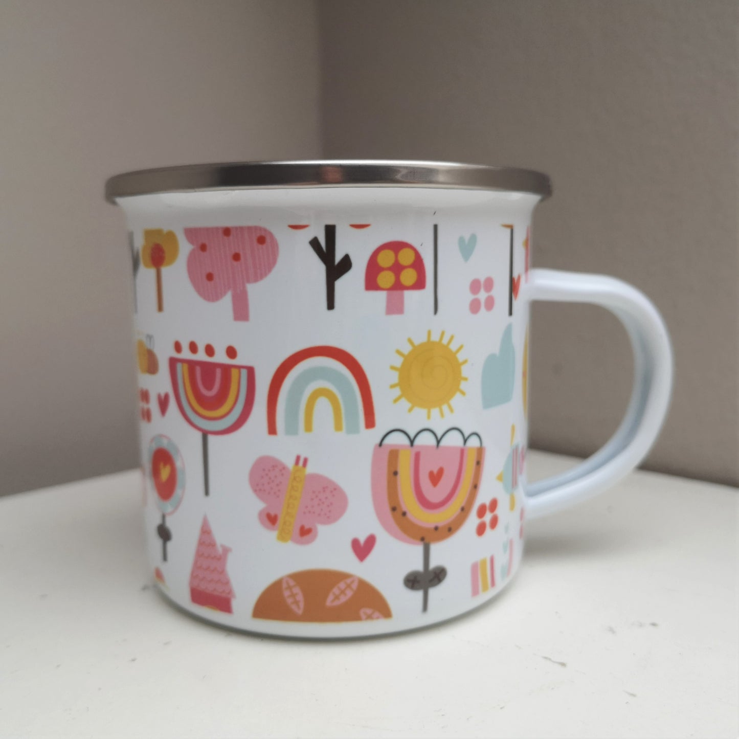 The back view of a White enamel mug with a stainless steel rim with a wraparound pink garden design and on the front a scalloped pink circle framing the owners name