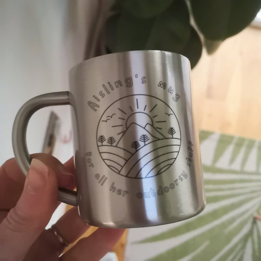 A personalised Stainless steel outdoor mug with a round mountains scene with a sun shining overheard, and the text on the mug is <NAME>'s mug for all his or her outdoorsy stuff