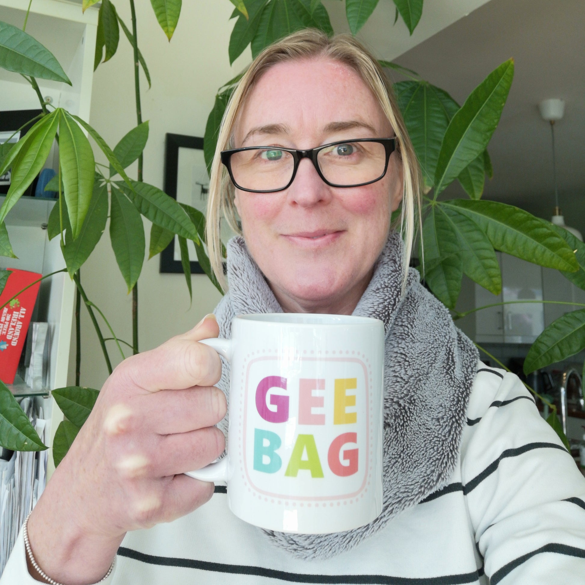A photo of me holding a white ceramic mug with GEEBAG written on the fron tin pastel lettering and spotty borders in pink