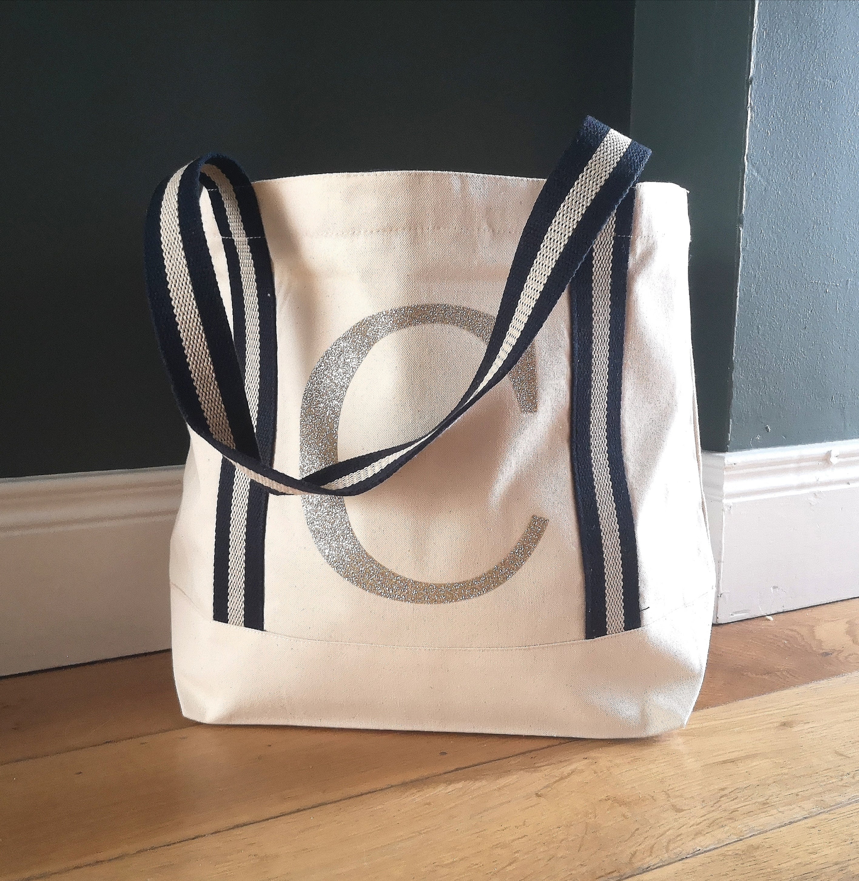 A Soft and sturdy natural cotton tote with navy and sparkly gold handles and a big personalised initial on the front in textured gold glitter