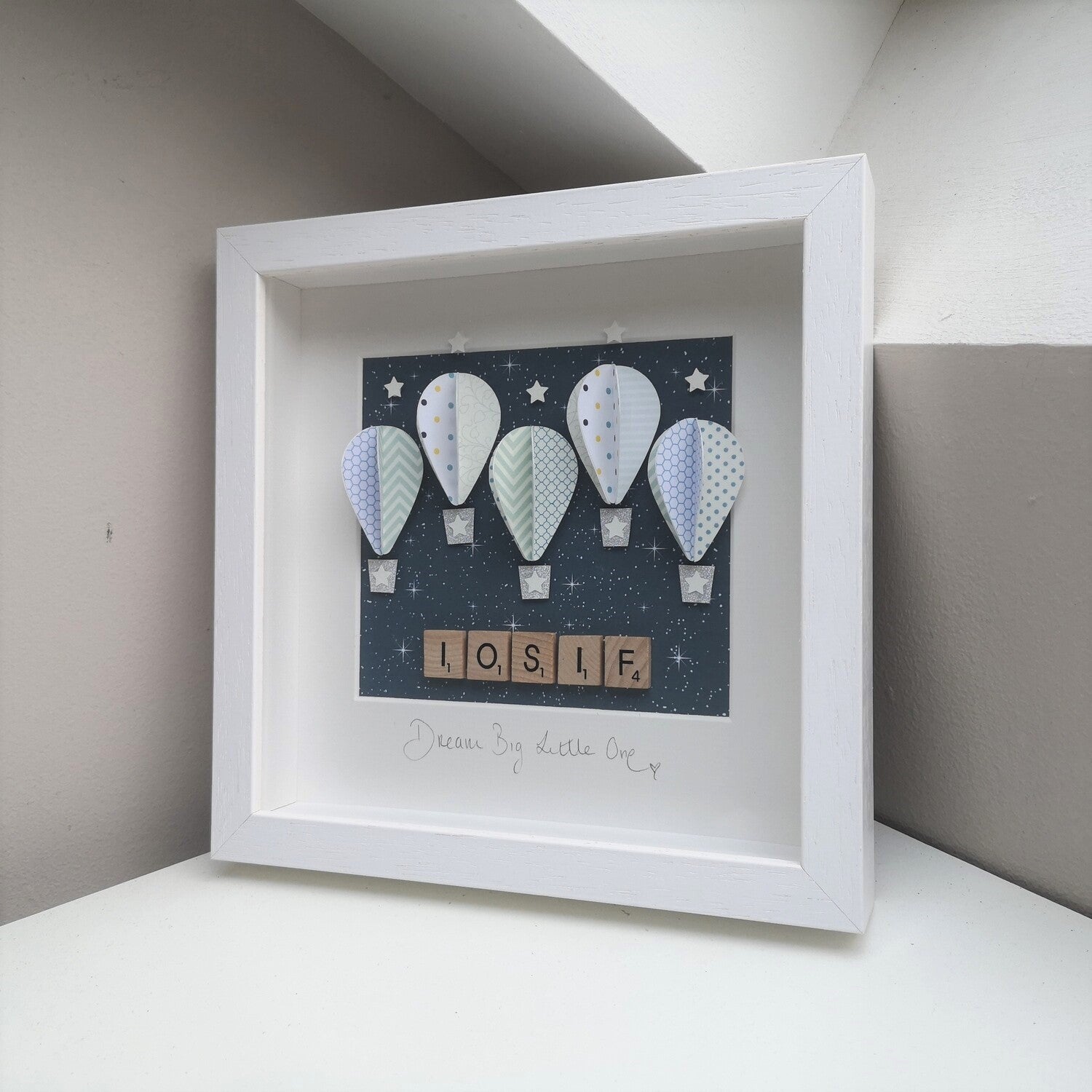 5 pastel blue patterned papercut balloons on a navy blue starry night backing, with a name in wooden scrabble tiles, in a handmade 25x25cm white deep box wooden frame.