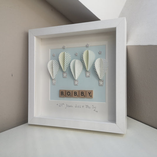 5 blue patterned papercut balloons on a pastel blue backing, with a name in wooden scrabble tiles, in a handmade 25x25cm white deep box wooden frame.