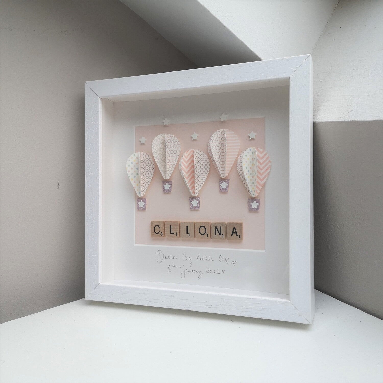5 pink patterned papercut balloons on a pastel pink backing, with a name in wooden scrabble tiles, in a handmade 25x25cm white deep box wooden frame.