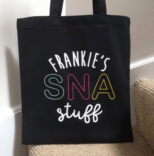 A personalised lightweight black cotton tote with <Name>'s SNA Stuff on it. SNA is in coloured pastel font.