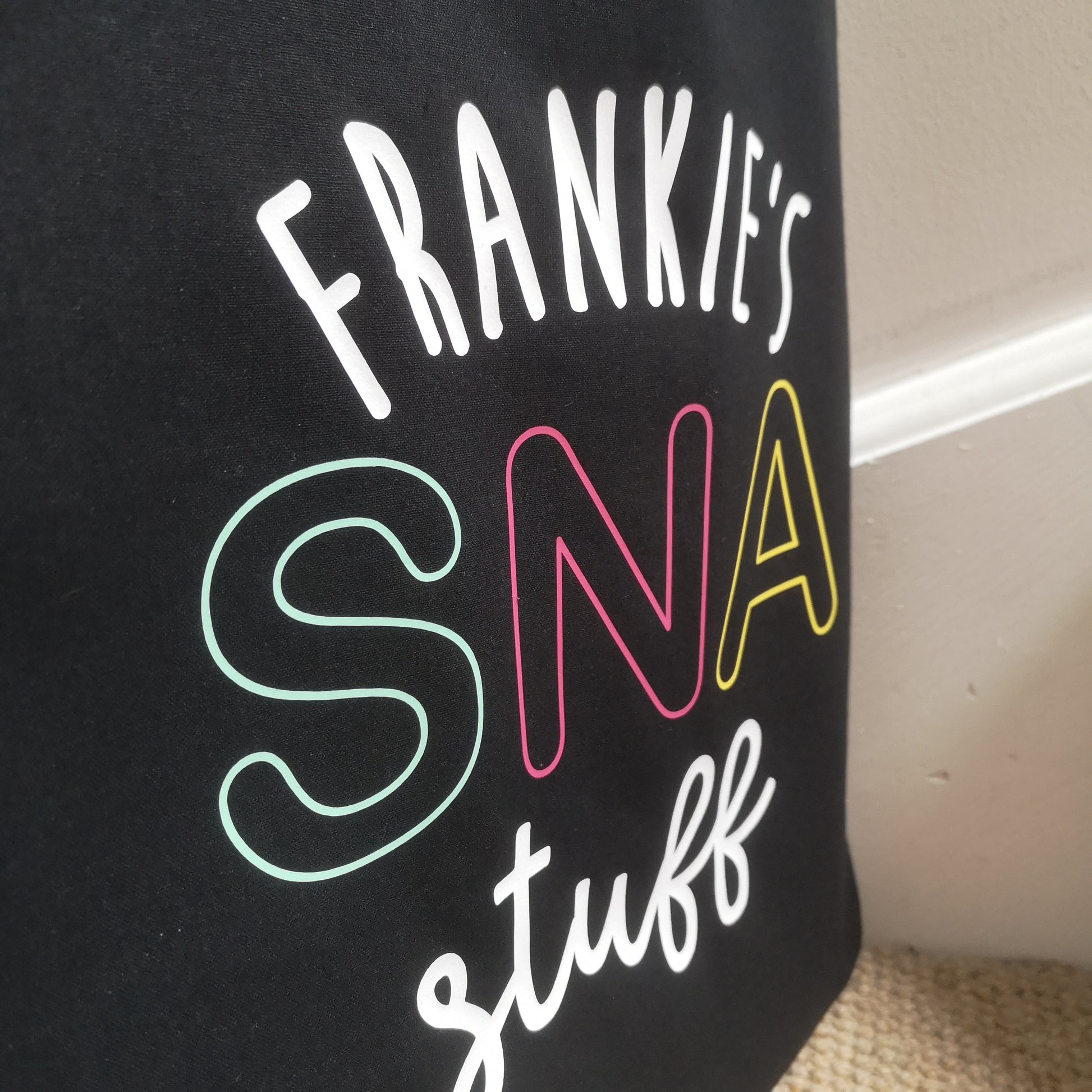 A close up photo of a personalised lightweight black cotton tote with <Name>'s SNA Stuff on it. SNA is in coloured pastel font.