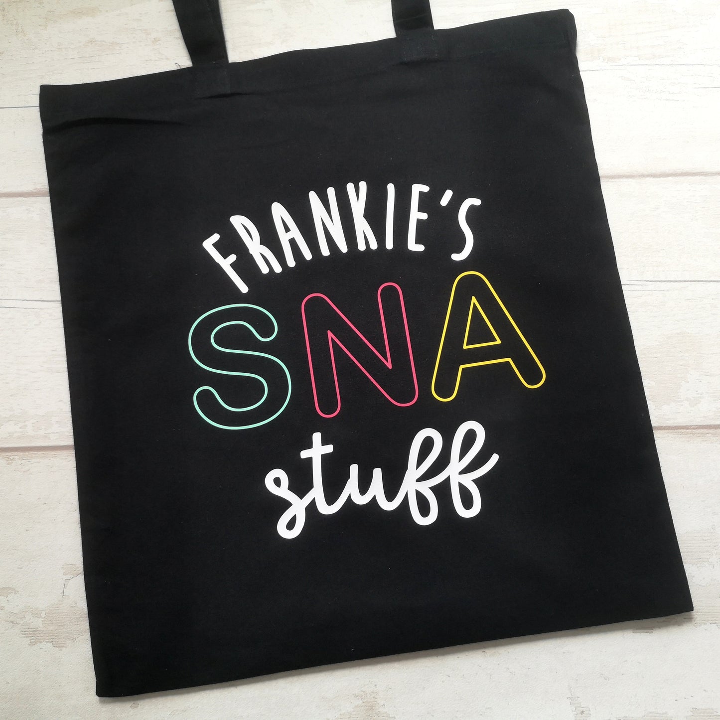 A different view of a  personalised lightweight black cotton tote with <Name>'s SNA Stuff on it. SNA is in coloured pastel font.