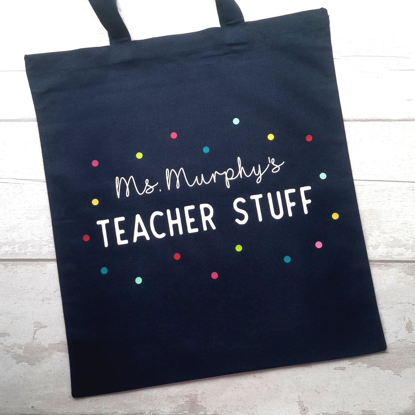 A personalised lightweight black cotton tote with <Name>'s TEACHER STUFF on it. The text is surrounded with multicoloured polkadots