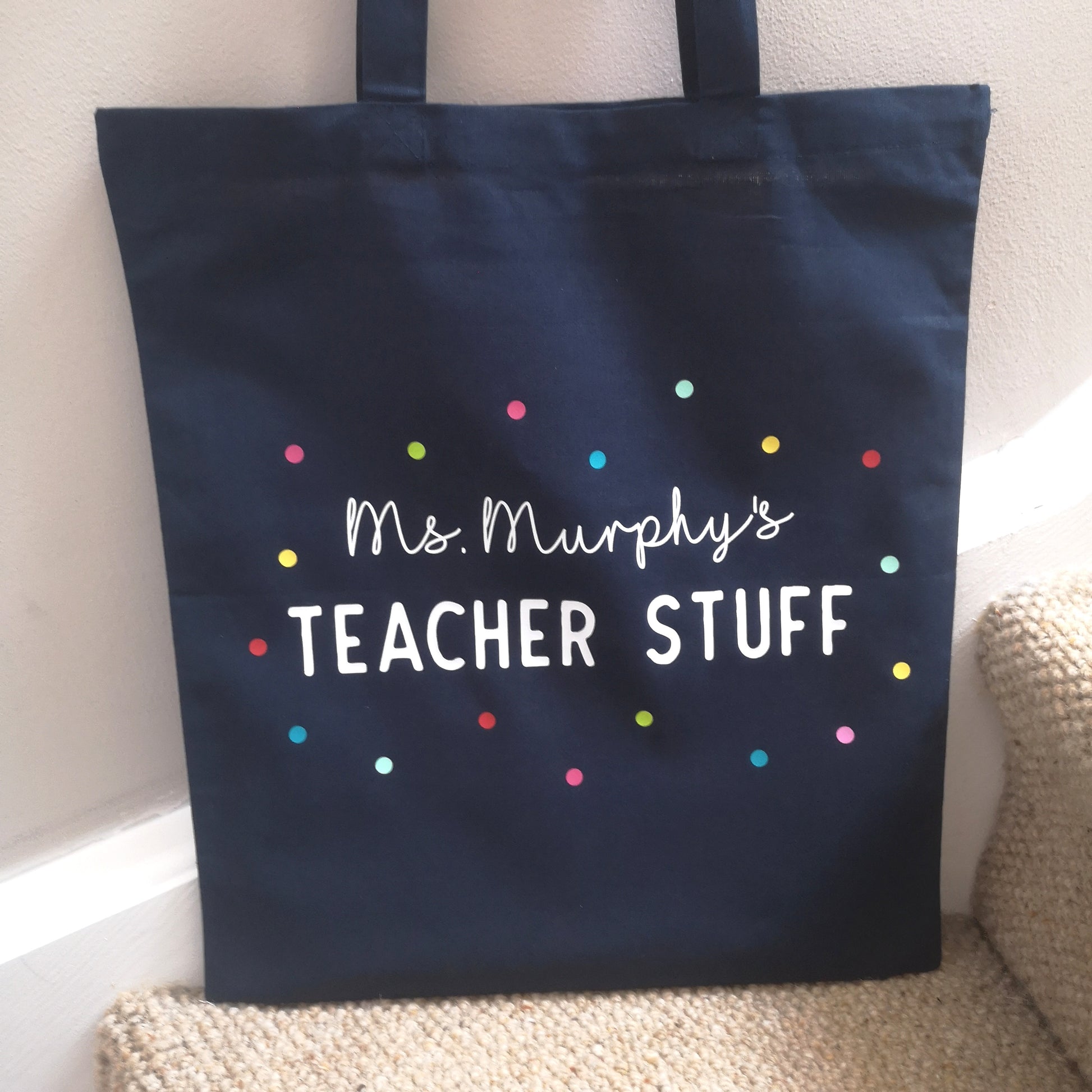 A personalised lightweight navy cotton tote with <Name>'s TEACHER STUFF on it. The text is surrounded with multicoloured polkadots