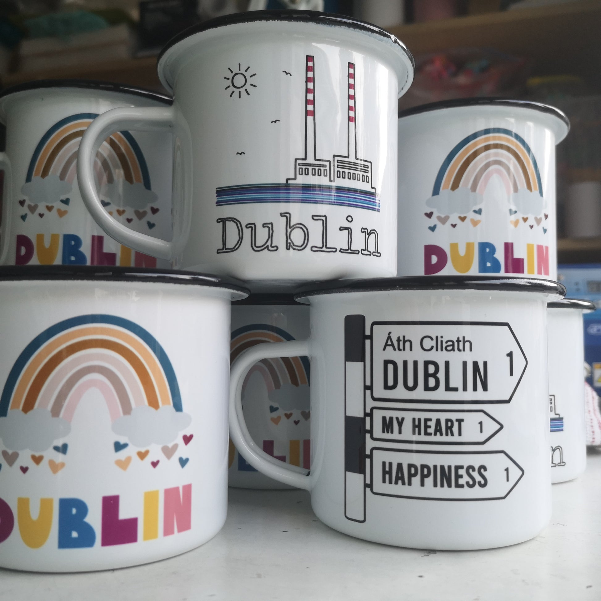 A photo of several Dublin enamel mugs, one with a rainbow, one with poolbeg towers on it and one with an old fashioned black and white road sign.