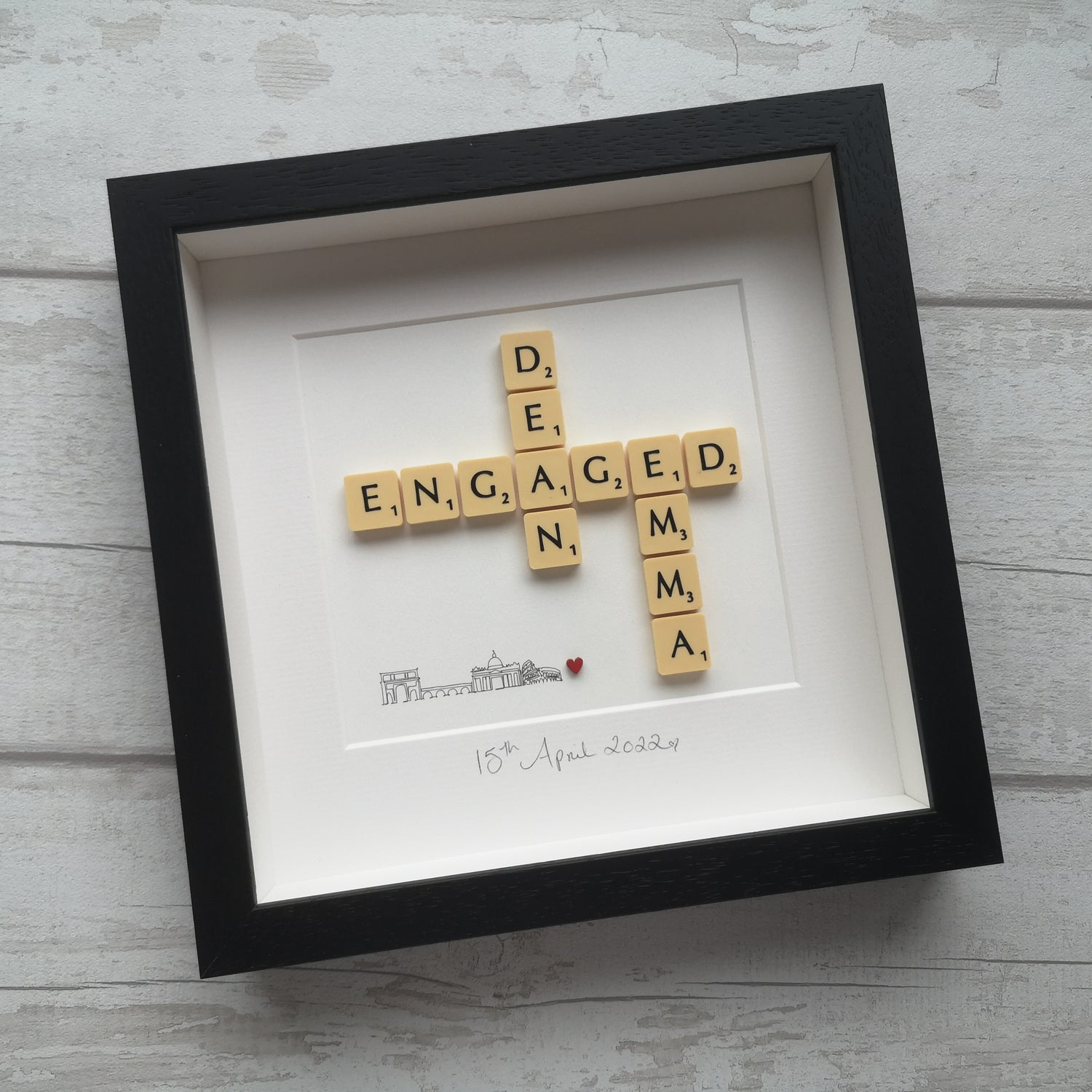 2 names and ENGAGED in vintage cream scrabble tiles on a off-white backing and a little red wooden love heart, with the date of their engagement written in pencil below. In a black handmade wooden 25x25cm deep box wooden frame.