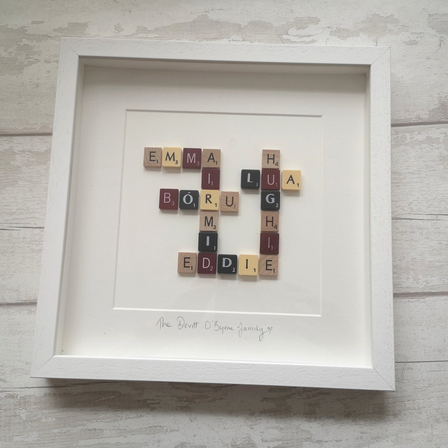 A white 30x30cm handmade wooden frame with mixed vintage and contemporary scrabble tiles, laid out like on a scrabble board.  Showing family names