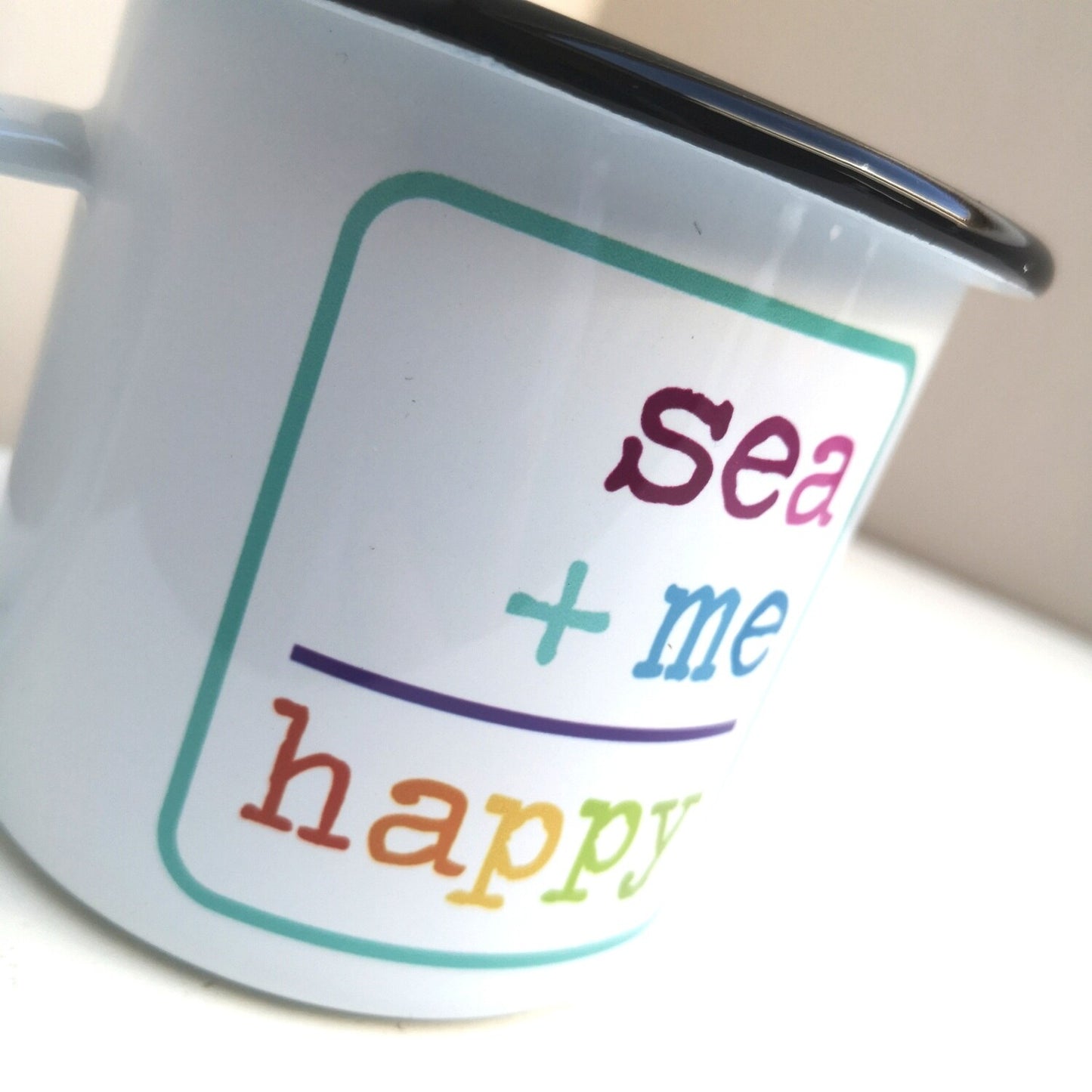 A close up of a White enamel mug with a black rim with the following on the front - SEA + ME = HAPPY in rainbow colours