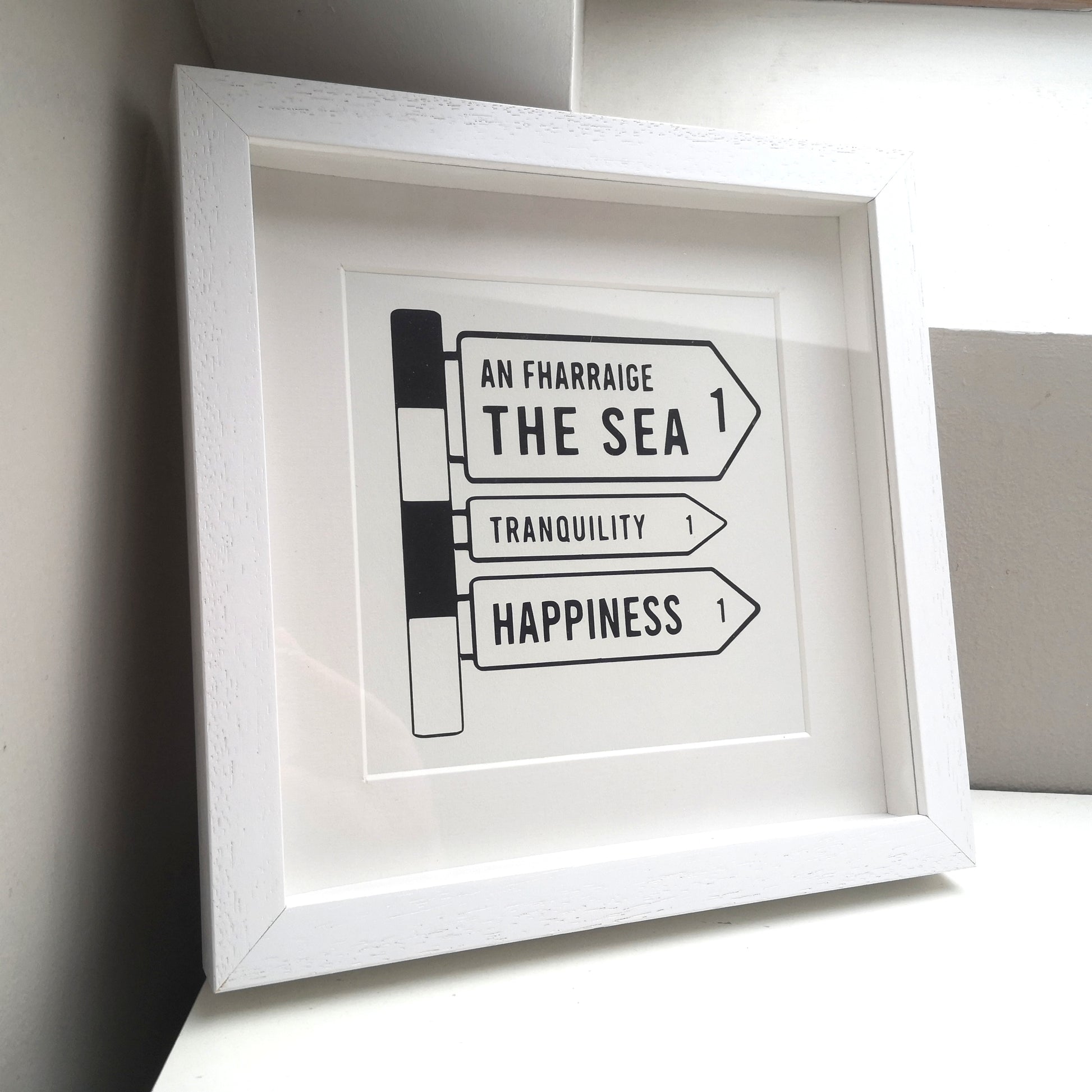 A 25x25cm deep box white wooden frame with a papercutting in it of an old Irish Roadsign pointing to the right with arrows for The Sea, Tranquility and Happiness