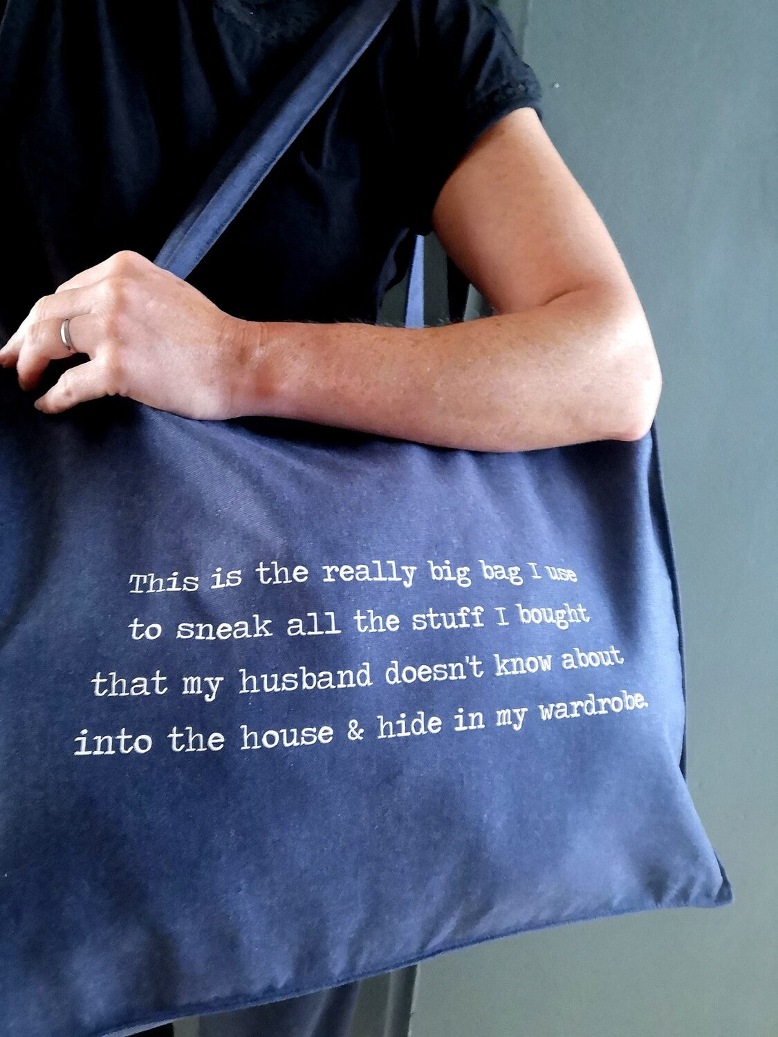 A woman carrying over her shoulder a large midnight blue organic cotton tote with the following text on it - This is the really big bag I use to sneak all the stuff I bought that my husband doesn't know about into the house & hide in my wardrobe.