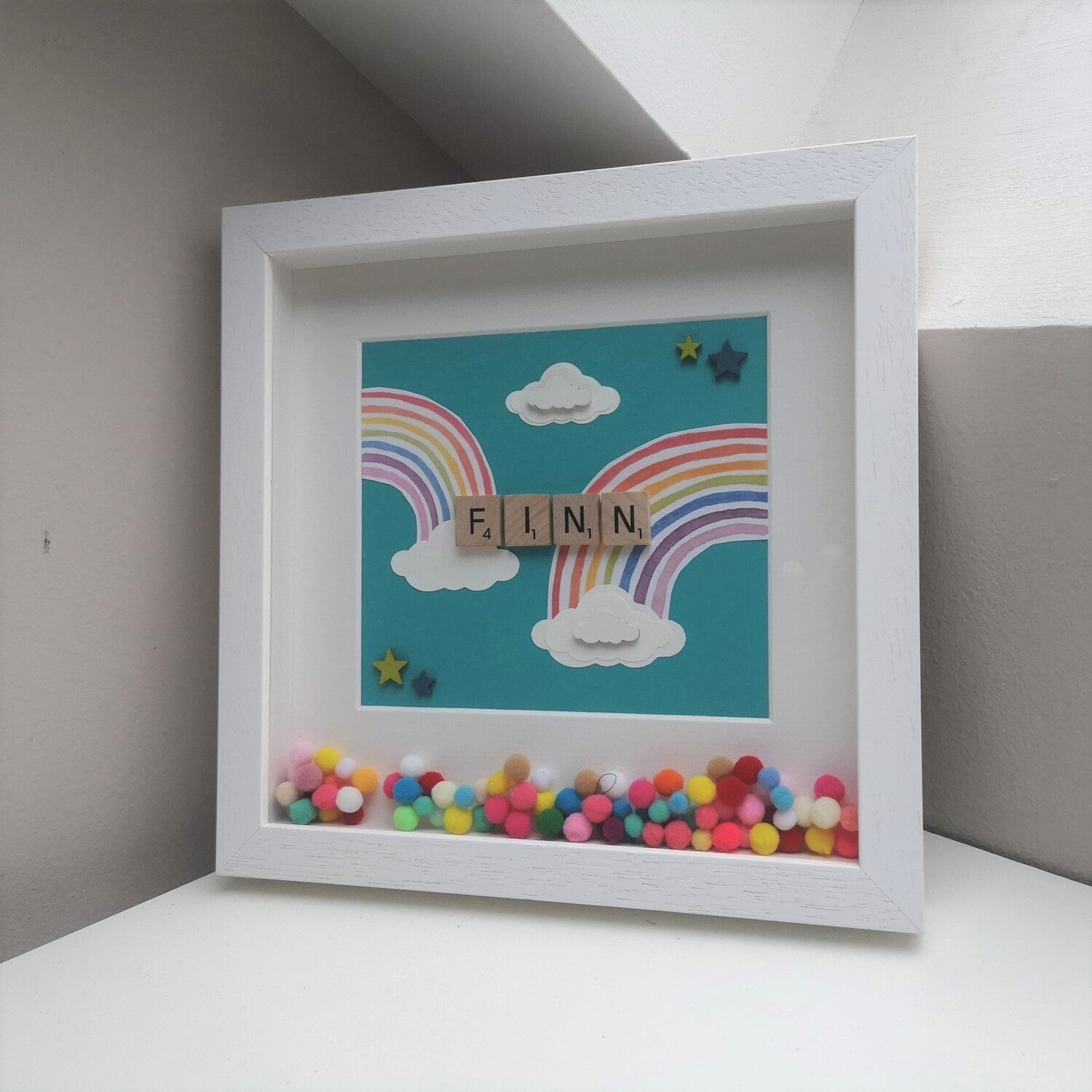 A name in wooden tiles on a  teal background with Papercut Rainbows and clouds, bright coloured pompoms and wooden clouds in a handmade 25x25cm deep box wooden frame.
