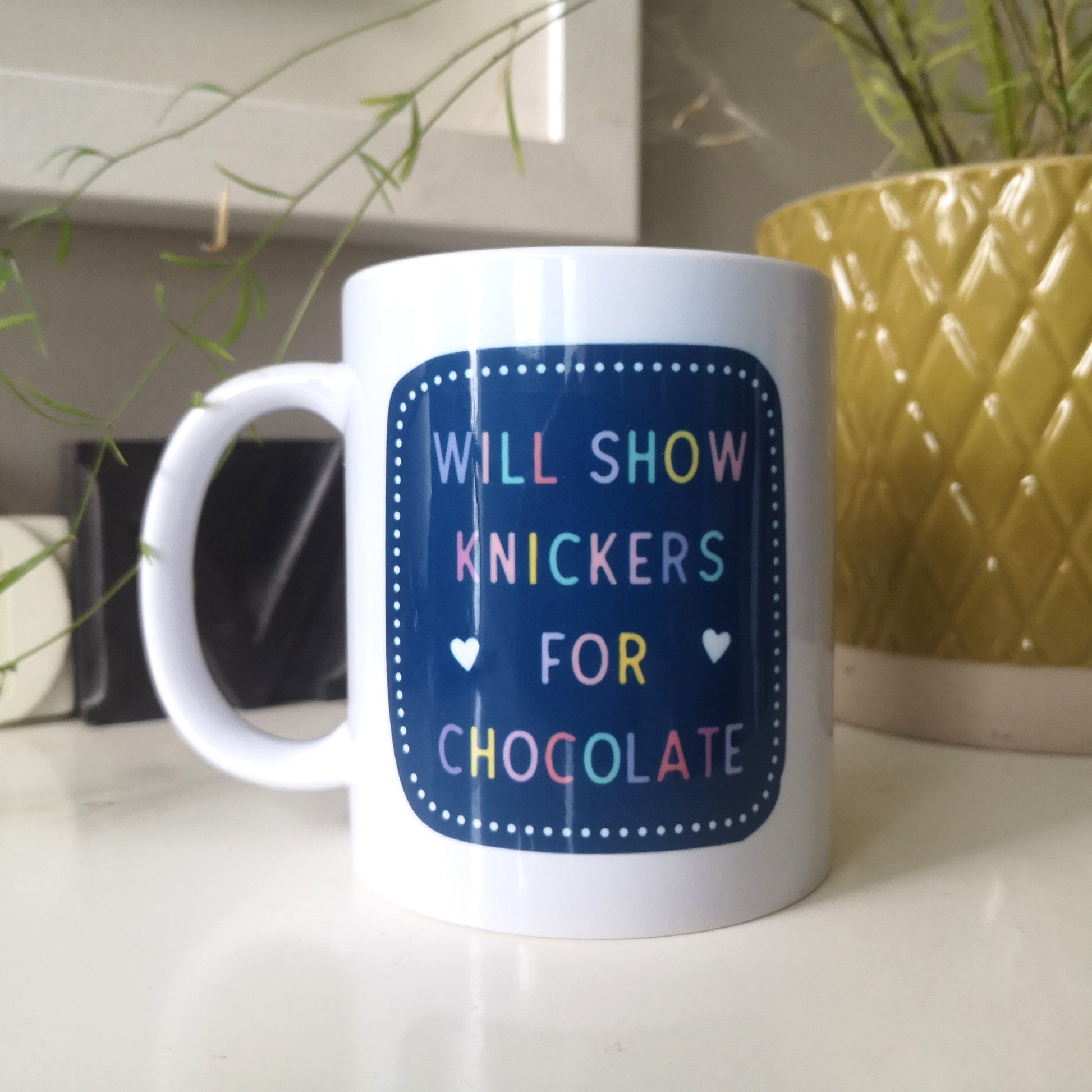 A ceramic mug with 'WILL SHOW KNICKERS FOR CHOCOLATE' in colourful writing on a dark blue background