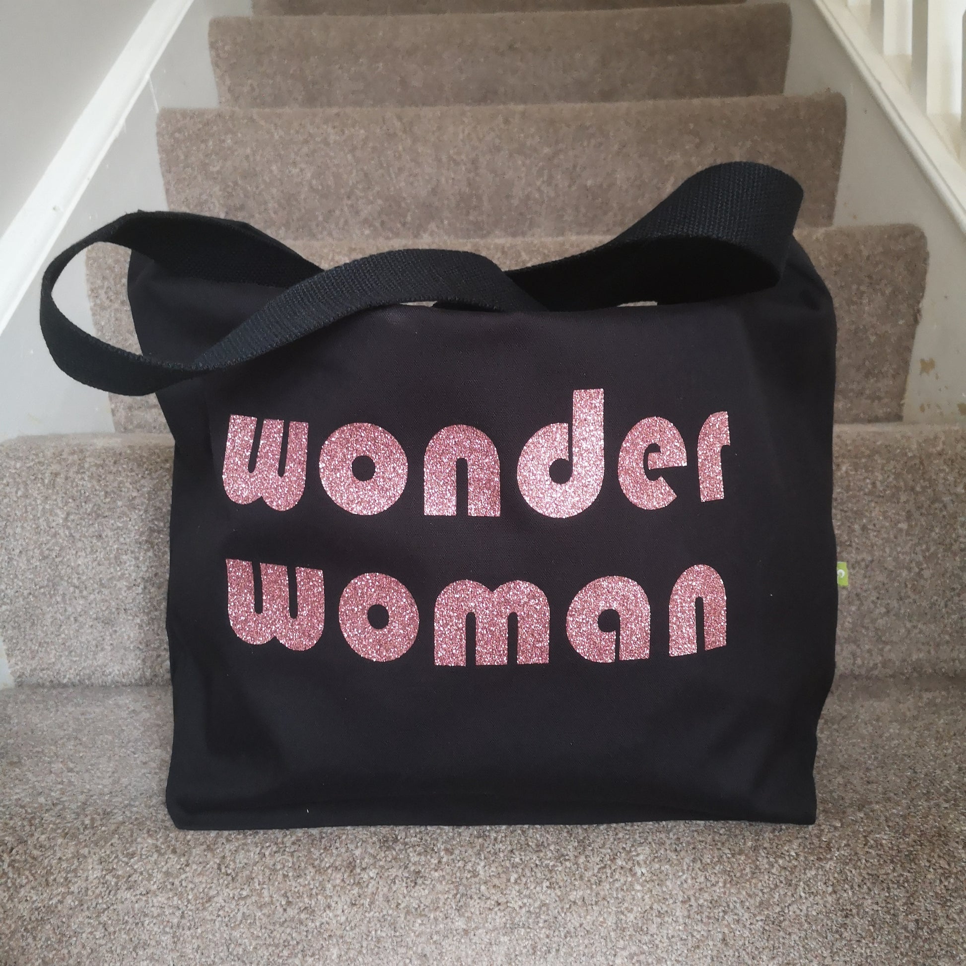 A black organic cotton tote with toggle closing with the following text on the front in glitter rose pink retro font - WONDER WOMAN