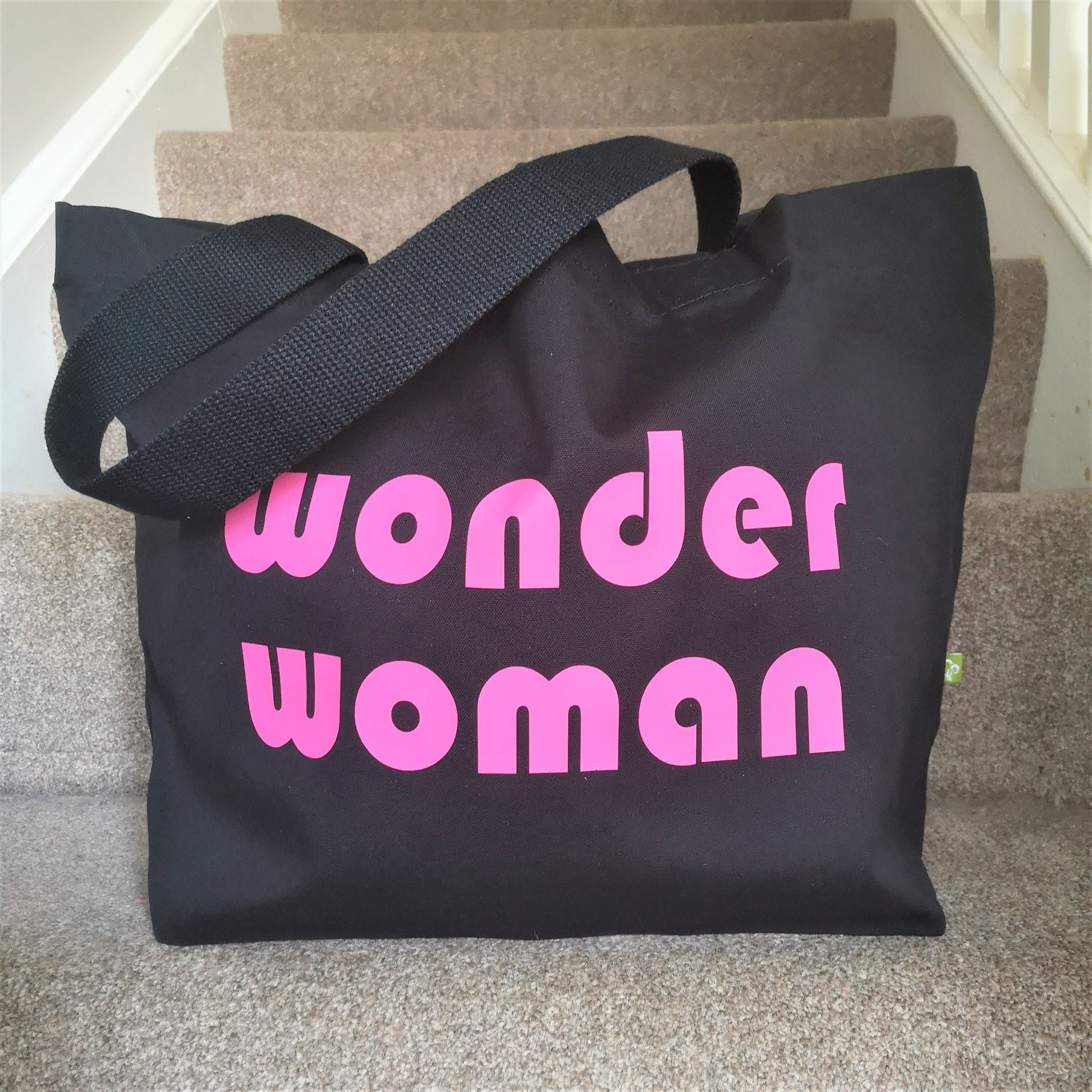 A black organic cotton tote with toggle closing with the following text on the front in neon pink retro font - WONDER WOMAN