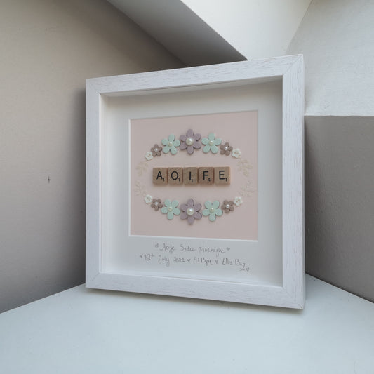 A pretty pastel wreath frame with a name in the middle in wooden letter tiles. The wreath is made with wooden flowers, pearls resin buttons and sparkly card leaves, in a handmade 25x25cm deep box wooden frame.